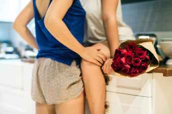 woman wearing blue tank top in front of woman sitting on kitchen cabinet with rose bouquet